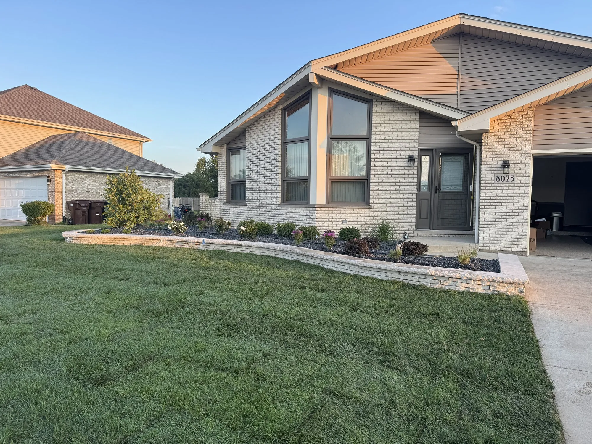 Golden-hour modern grey-brick home with a curved stacked-stone landscape bed by Rodak Landscaping
